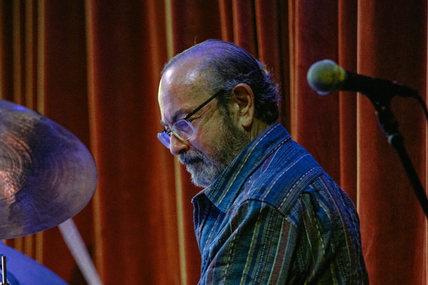 Peter Kogan seated with drumsticks in hand, playing drum set, a red curtain behind him at Icehouse